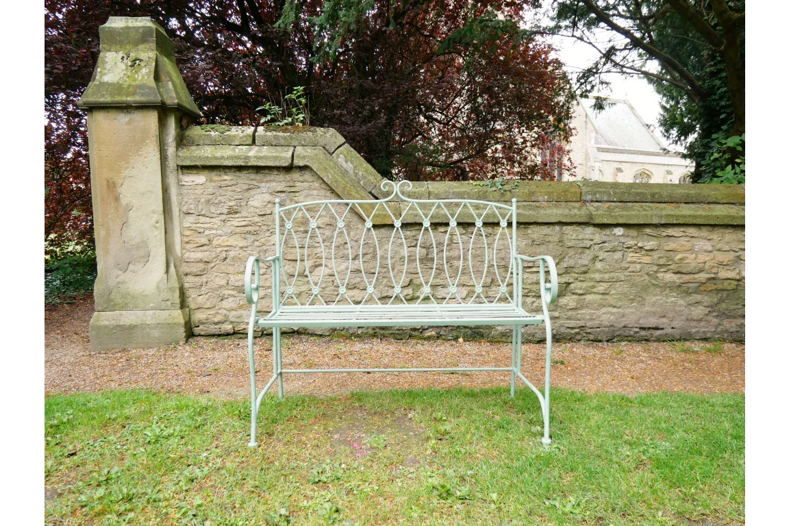 Green metal garden bench with decorative backrest near stone wall and trees