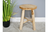 Round natural wood stool with three legs and cross braces, angled view on carpeted floor beside green plant in black pot.