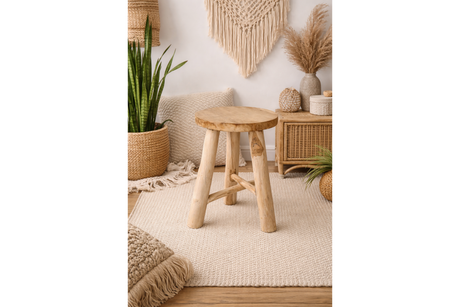 Round wooden stool with three legs in a beige, bohemian-style room setting with plants and woven decor.