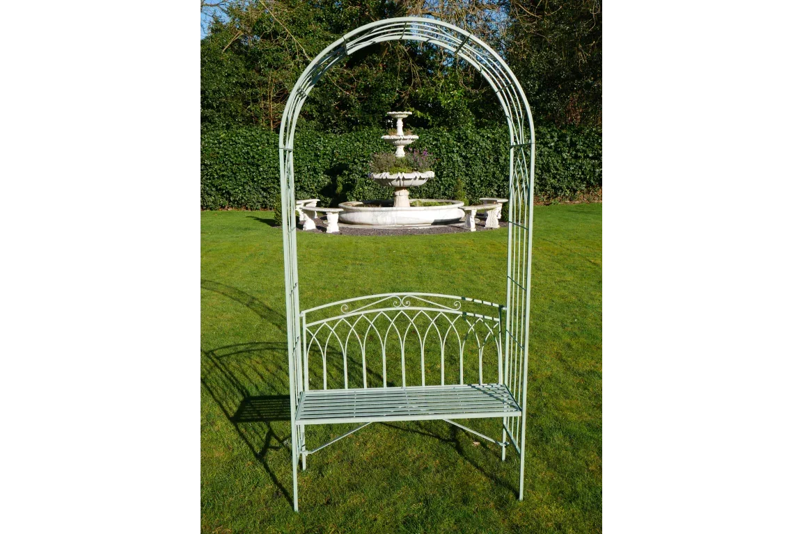 Green metal garden bench with arch frame on lawn, fountain in background