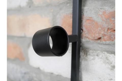 Close-up of a black metal cylindrical wine bottle holder attached to a vertical bar against a brick wall background.