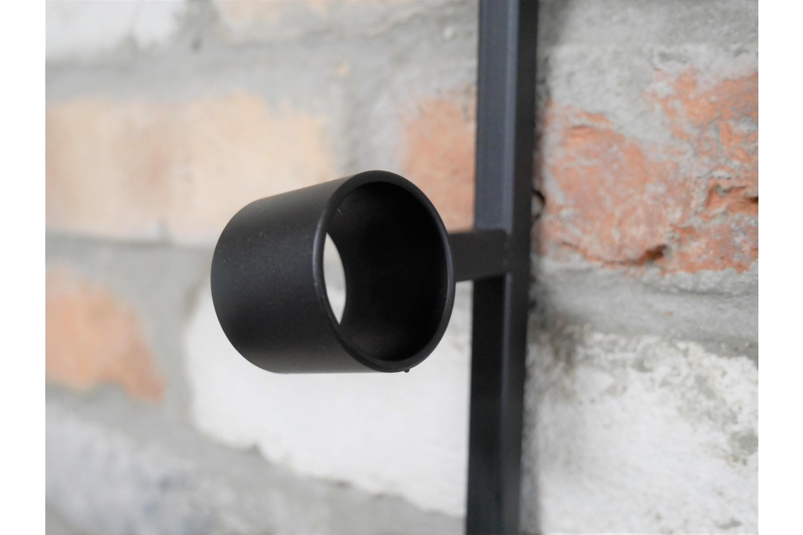 Close-up of a black metal cylindrical wine bottle holder attached to a vertical bar against a brick wall background.