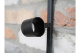 Close-up of a black metal cylindrical wine bottle holder attached to a vertical bar against a brick wall background.