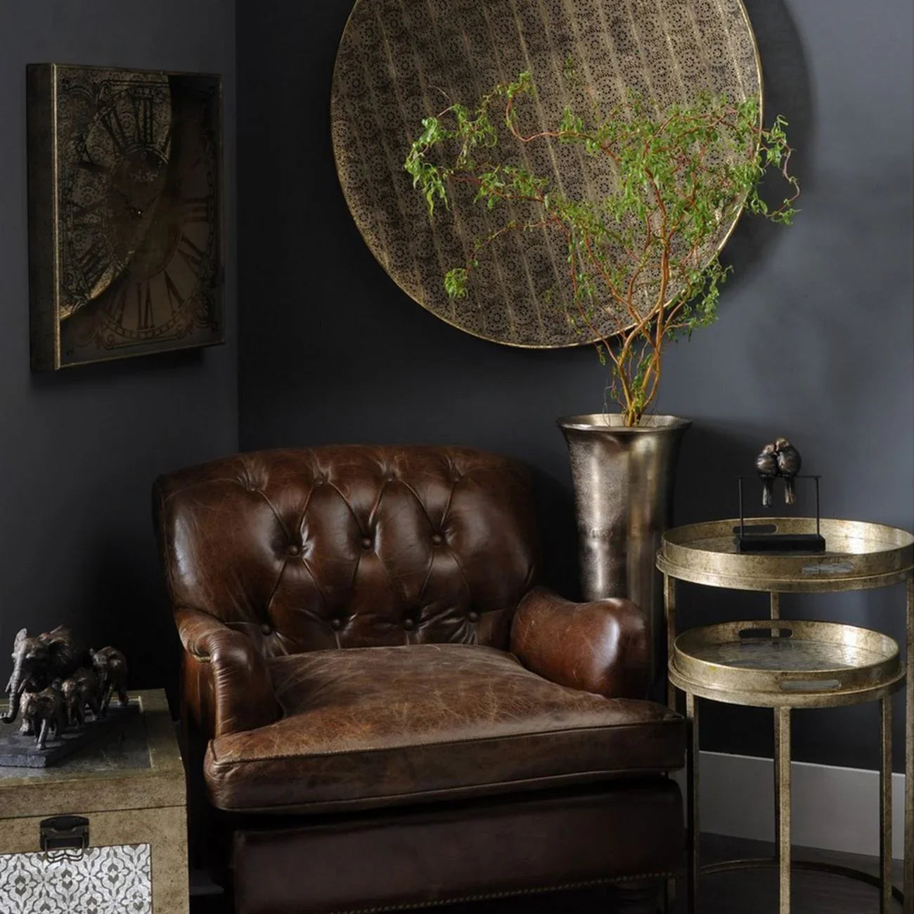 Antique gold tray tables beside a brown leather armchair in a dark, elegant room.