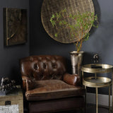 Antique gold tray tables beside a brown leather armchair in a dark, elegant room.