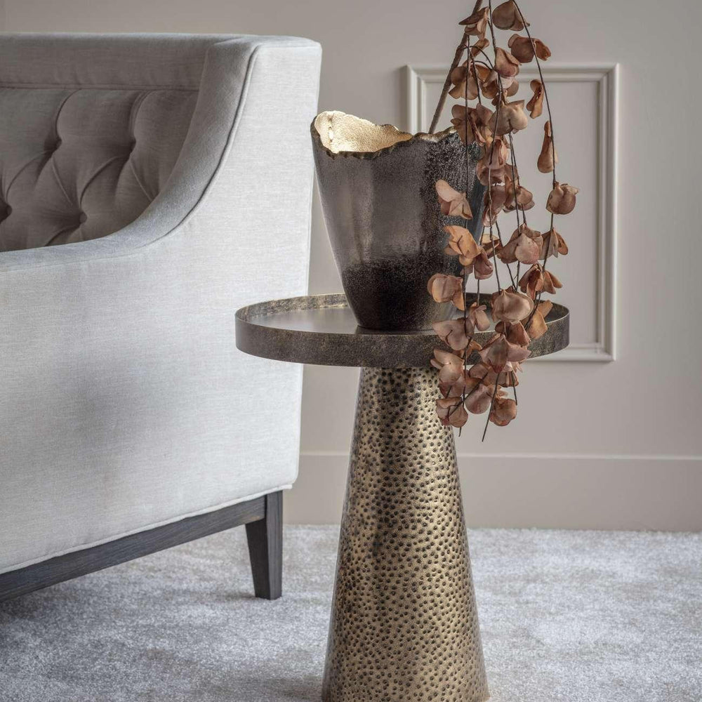 Rustic gold side table with textured base beside a grey sofa.