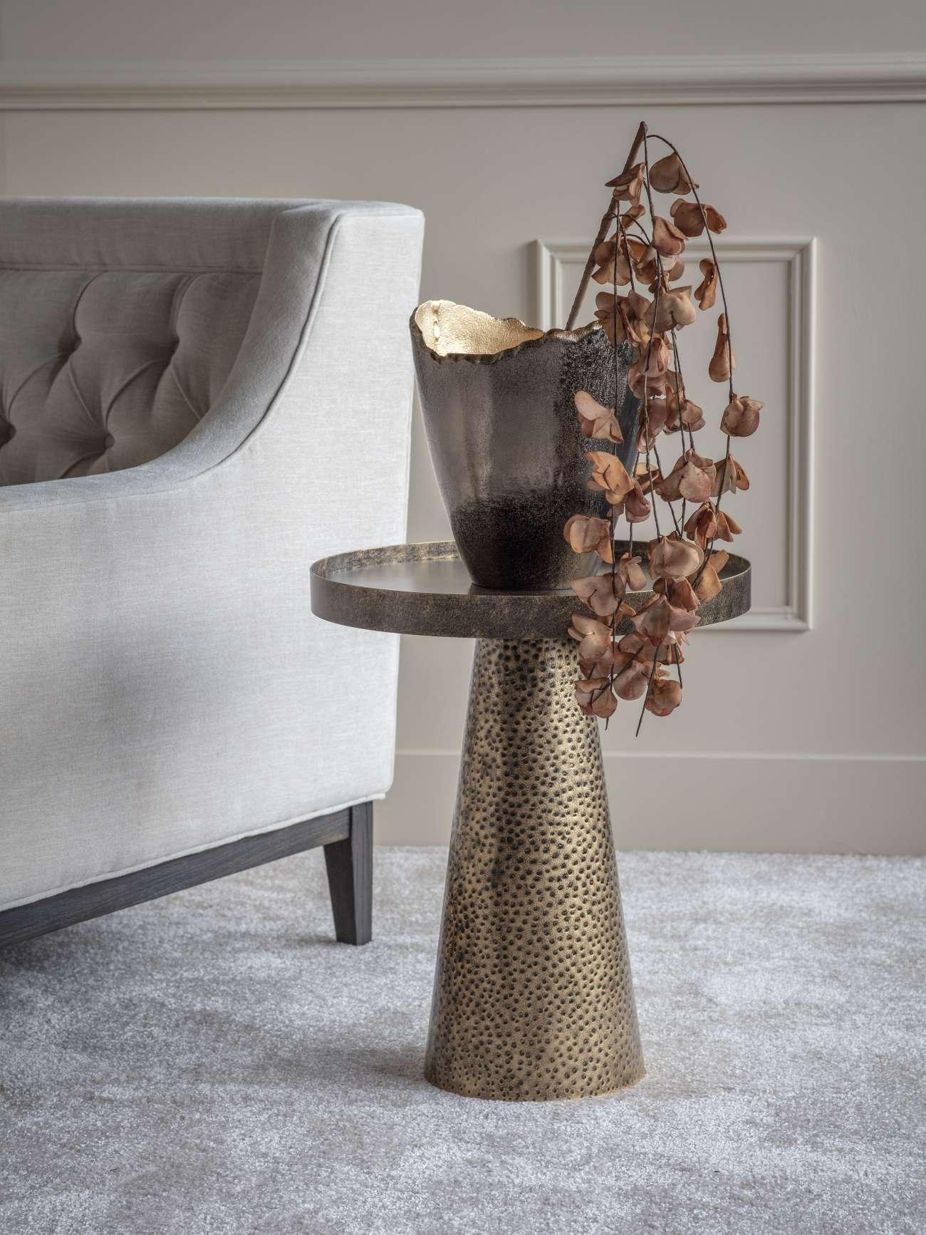 Rustic gold side table with textured base beside a grey sofa.