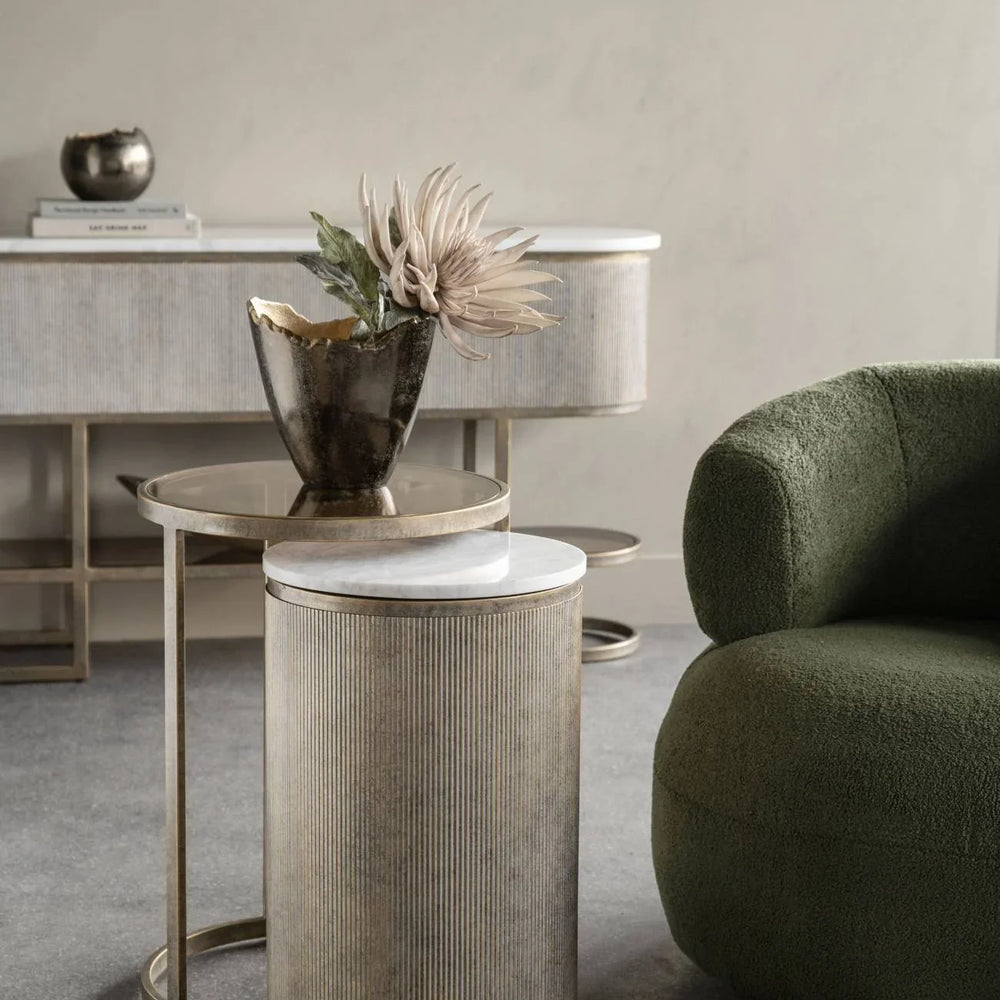 Aged gold nesting tables with a vase beside a green armchair in a stylish room.