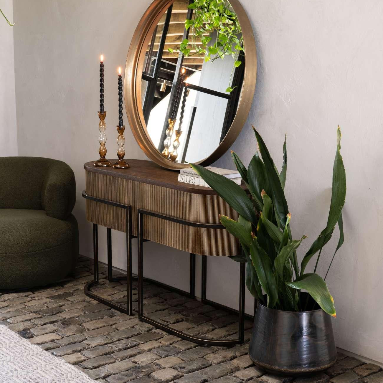 Gold console table with round mirror, styled with plants and candles on cobblestone floor.