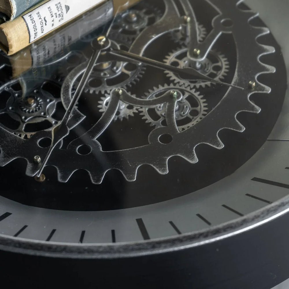 Black nickel clock with visible moving cogs and books on glass surface.