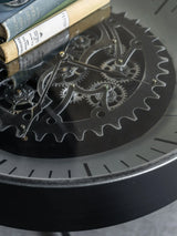 Black nickel clock with visible moving cogs and books on glass surface.