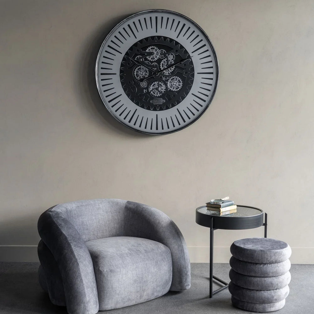 Round wall clock with visible cogs above a grey chair and side table in a modern room.