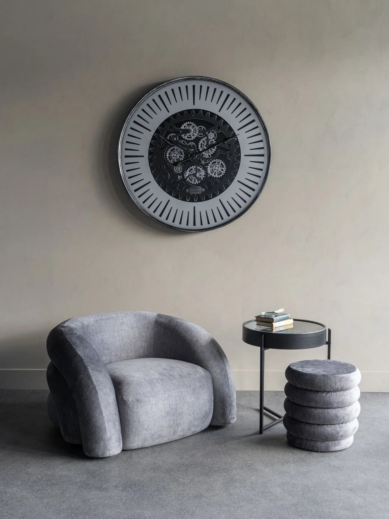 Round wall clock with visible cogs above a grey chair and side table in a modern room.