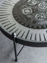 Black metal clock table with intricate moving gears and Roman numerals.
