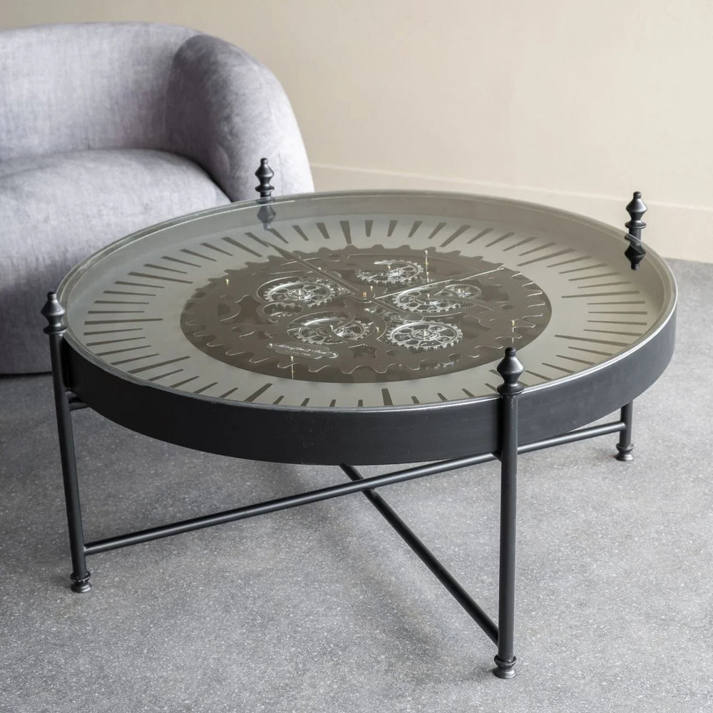Round glass coffee table with visible cogs, set on a grey floor near a sofa.