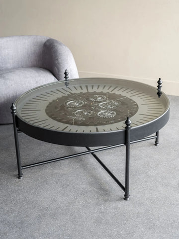 Round glass coffee table with visible cogs, set on a grey floor near a sofa.