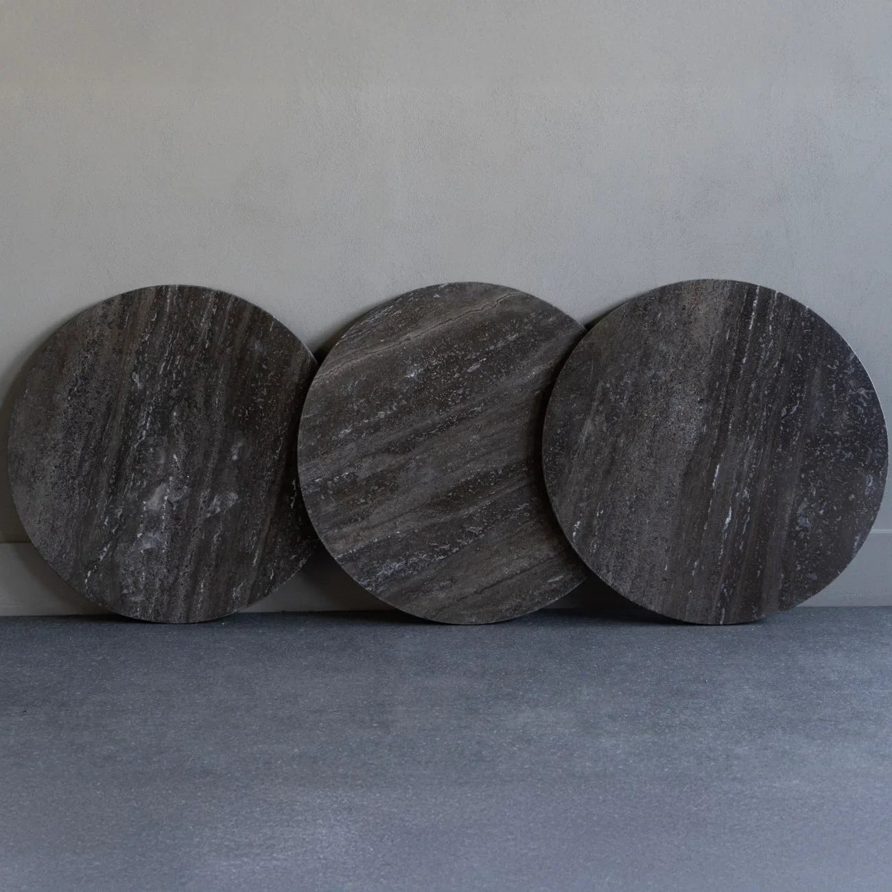 Three dark travertine round tabletops leaning against a wall.