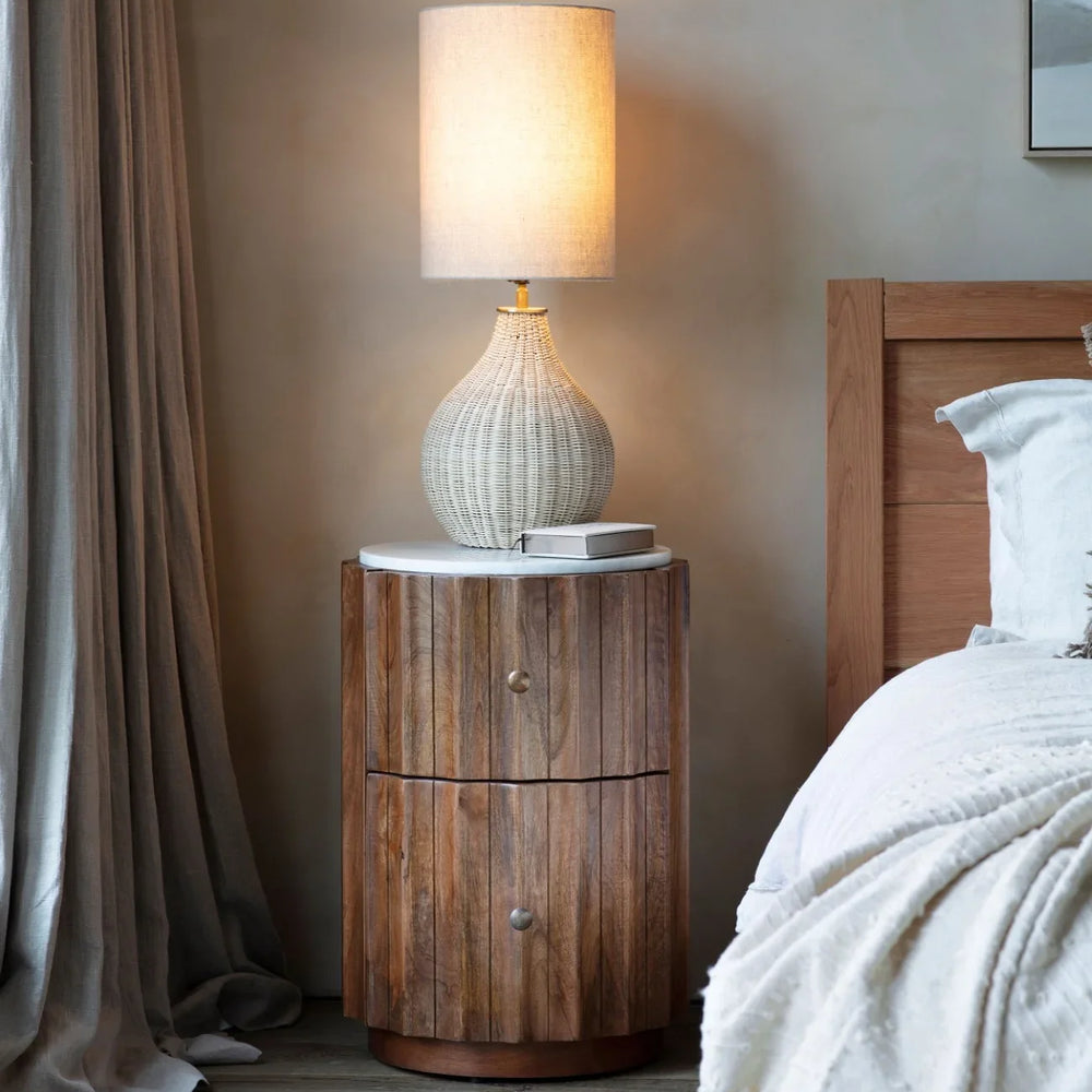 Wooden bedside table with marble top beside bed and lamp in cozy bedroom.