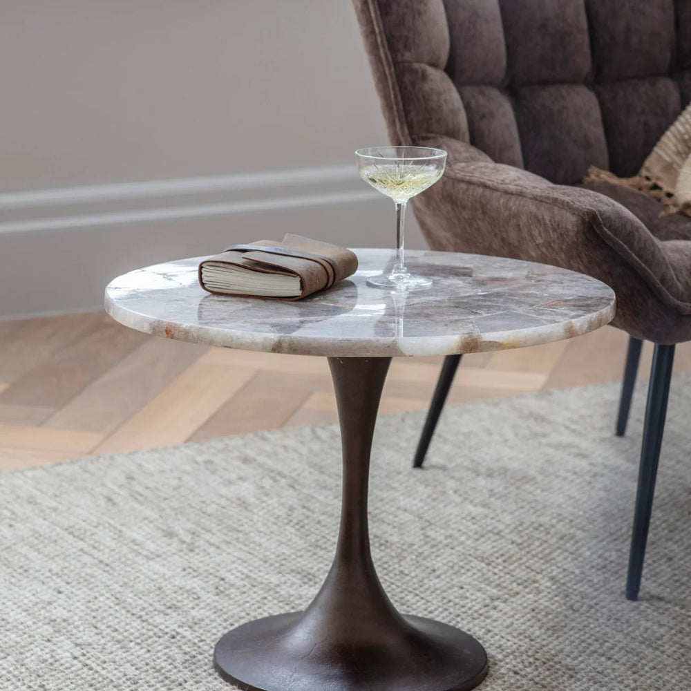 Round stone side table with a glass and book, next to a plush brown chair.