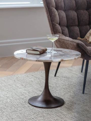 Round stone side table with a glass and book, next to a plush brown chair.