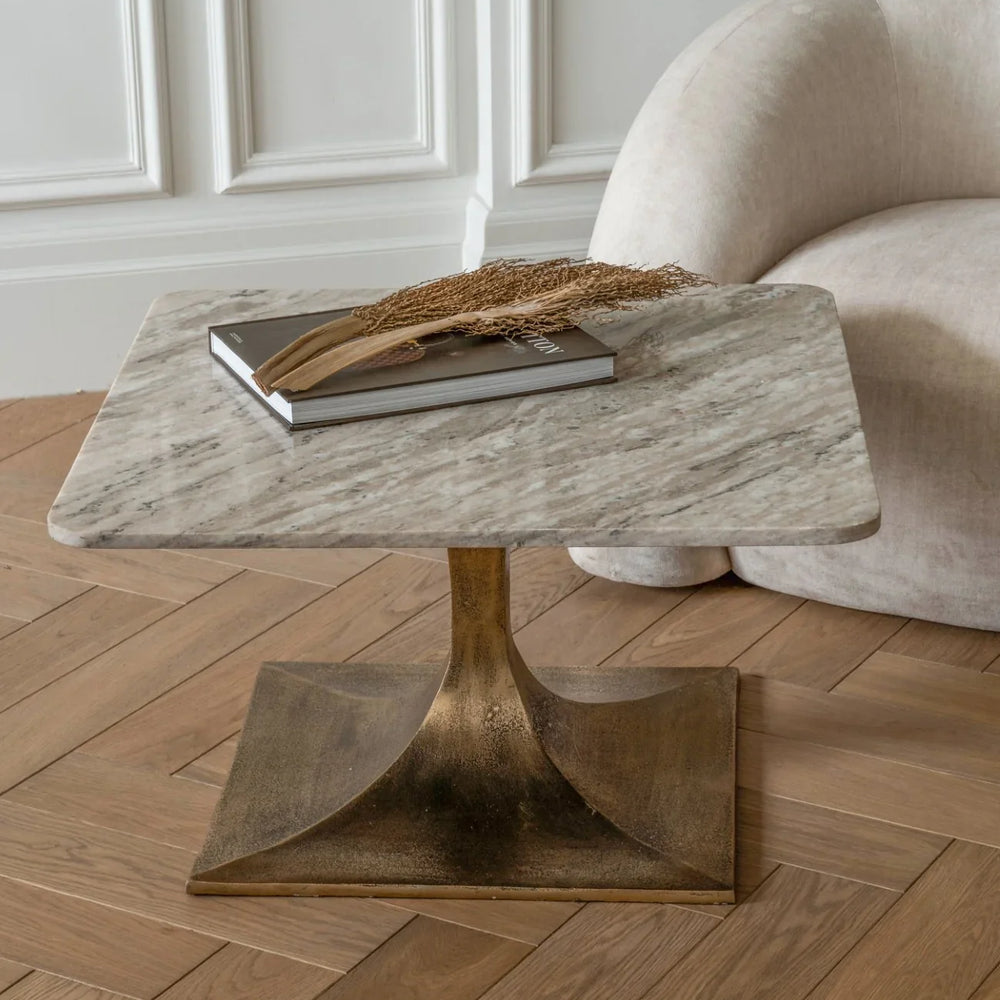 Square marble table with gold base on wooden floor, styled with book and dried foliage.