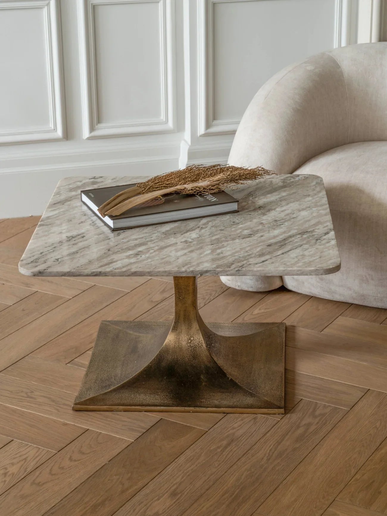 Square marble table with gold base on wooden floor, styled with book and dried foliage.