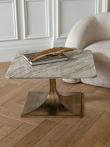 Square marble table with gold base on wooden floor, styled with book and dried foliage.