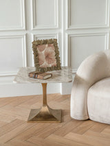 Marble side table with brass base beside a beige sofa and decorative frame.