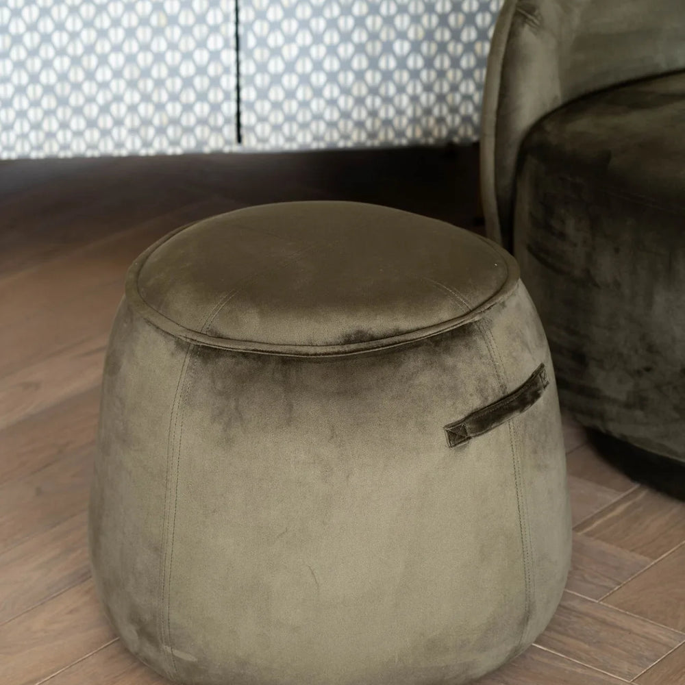Green velvet stool on wooden floor with patterned curtain backdrop.