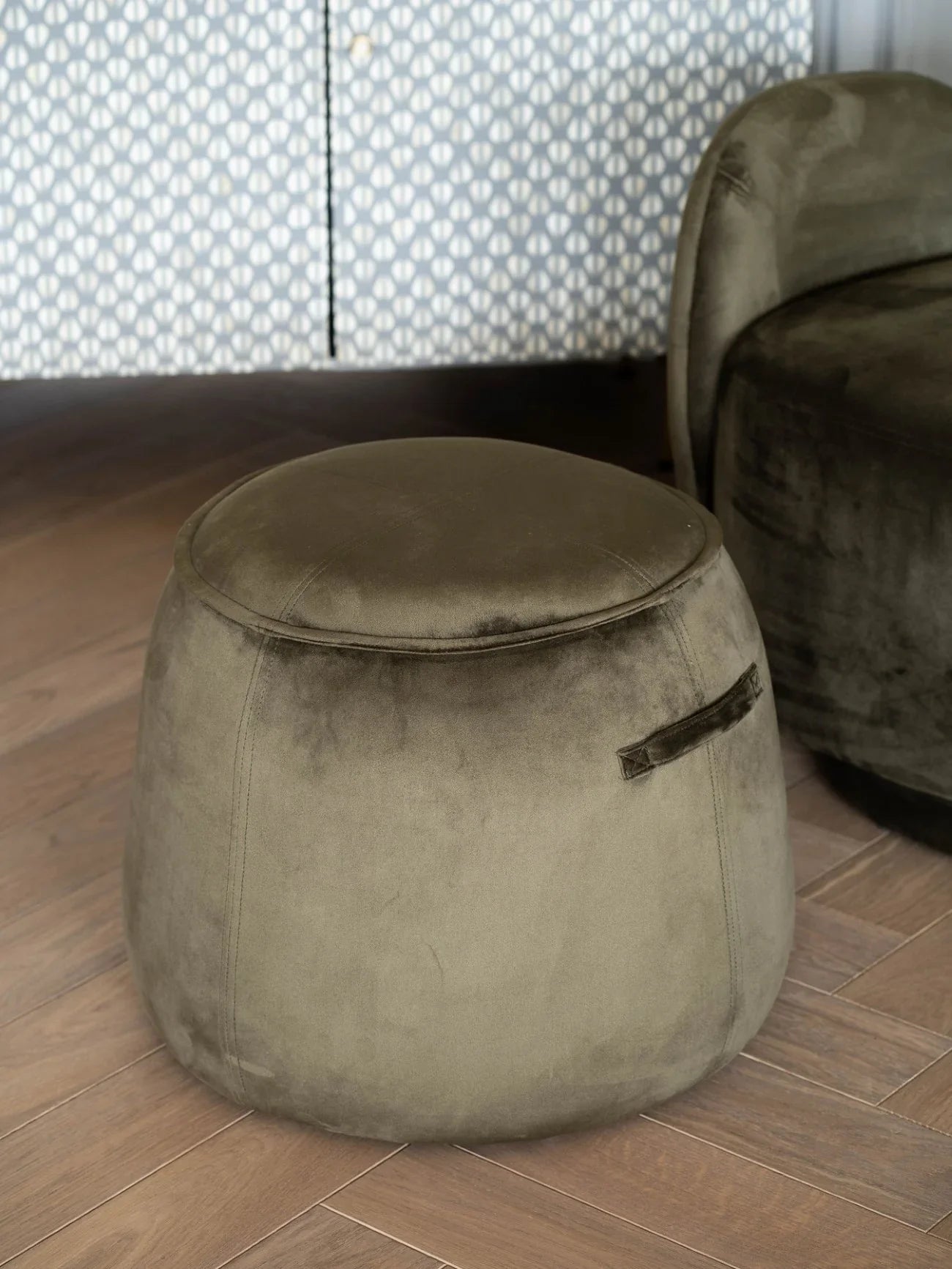 Green velvet stool on wooden floor with patterned curtain backdrop.