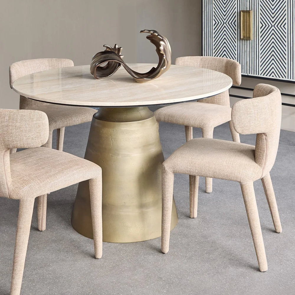 Beige dining chairs around a round travertine table with a bronze sculpture centerpiece.