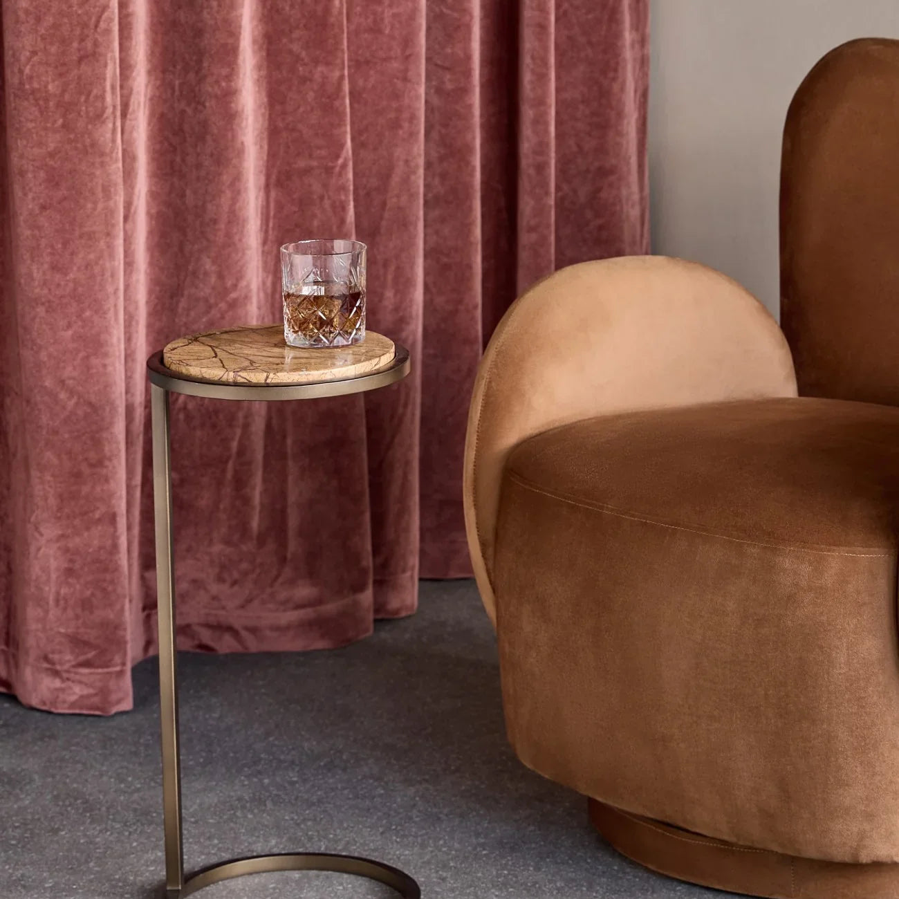 Marble-topped side table with gold frame beside a plush brown chair and pink curtains.