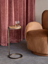 Marble-topped side table with gold frame beside a plush brown chair and pink curtains.