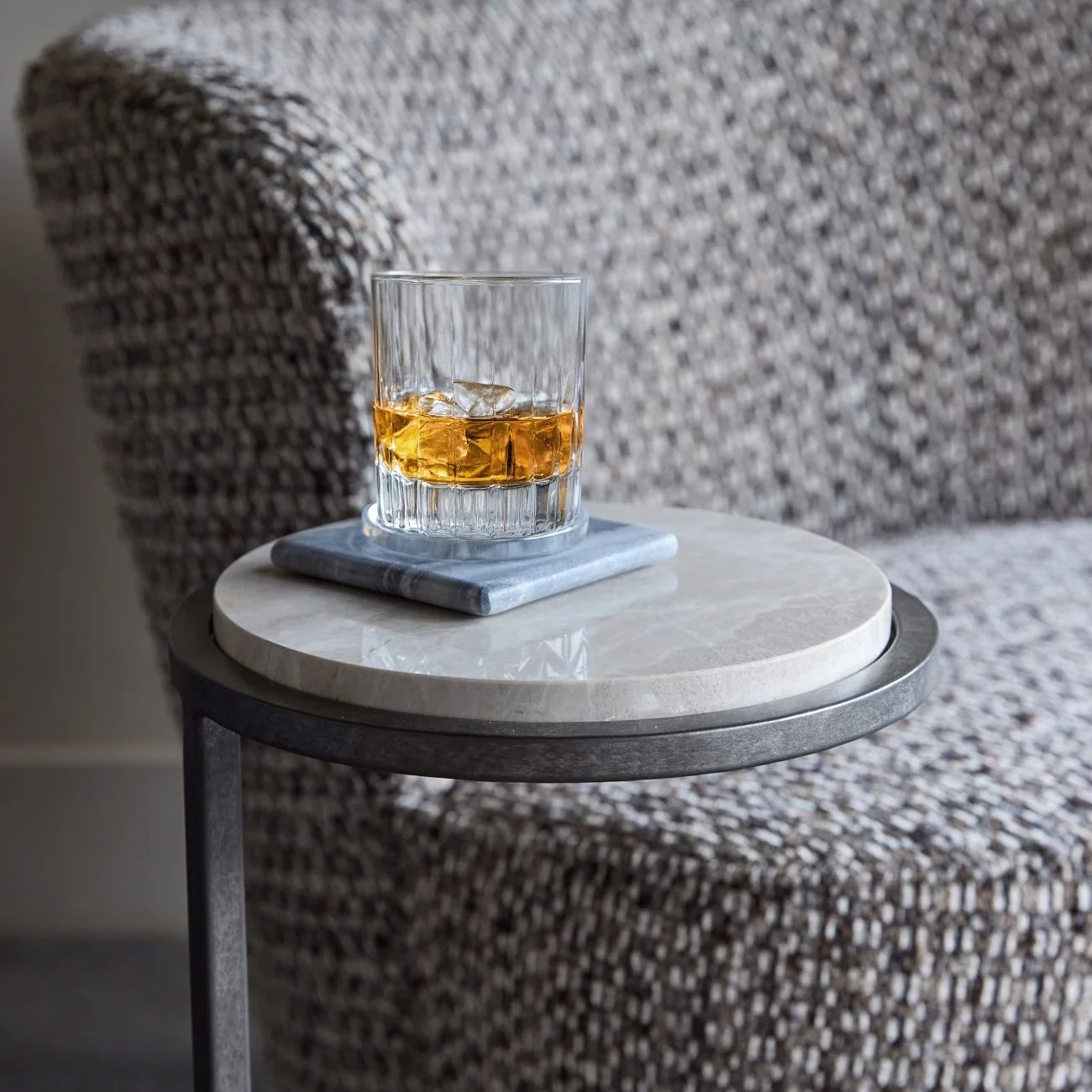Glass of whiskey on grey marble table beside textured armchair.