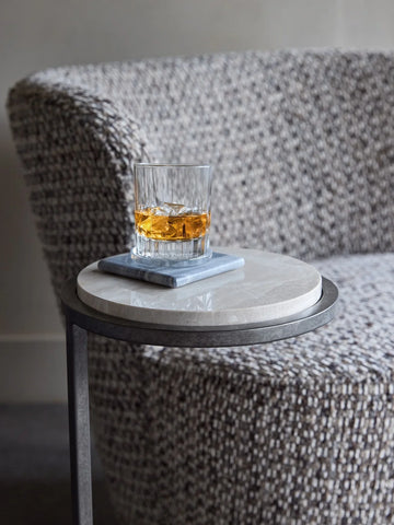 Glass of whiskey on grey marble table beside textured armchair.