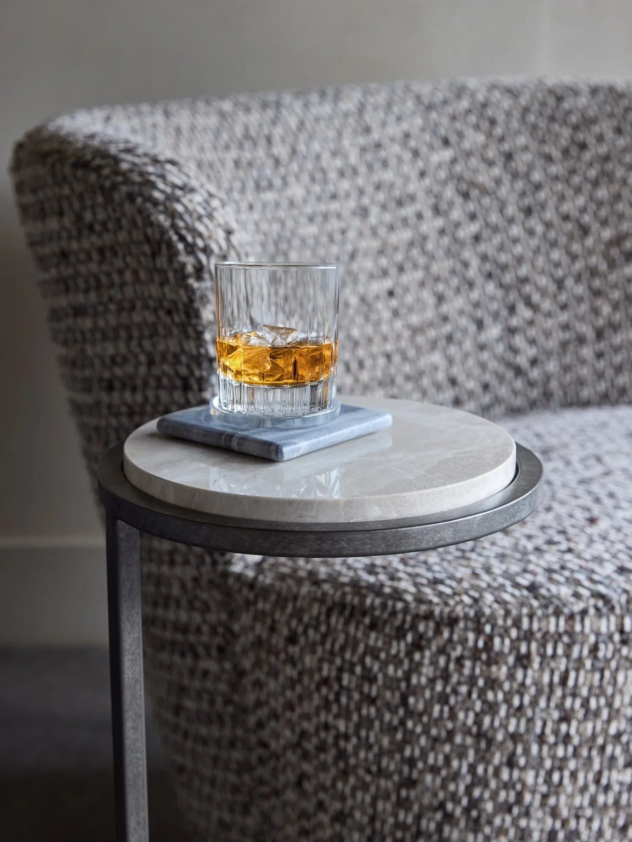 Glass of whiskey on grey marble table beside textured armchair.
