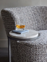 Glass of whiskey on grey marble table beside textured armchair.