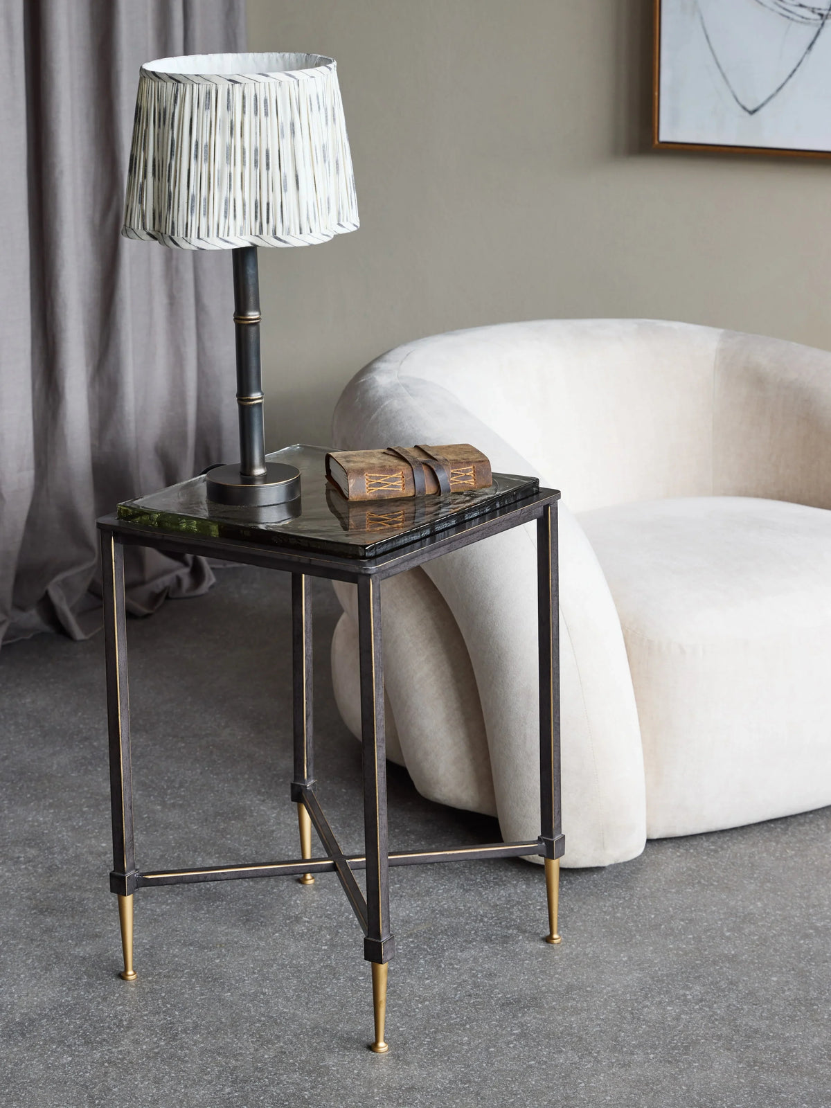 Black metal side table with glass top, styled with lamp and book in modern living room.