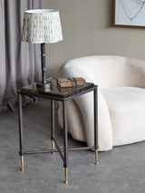 Black metal side table with glass top, styled with lamp and book in modern living room.