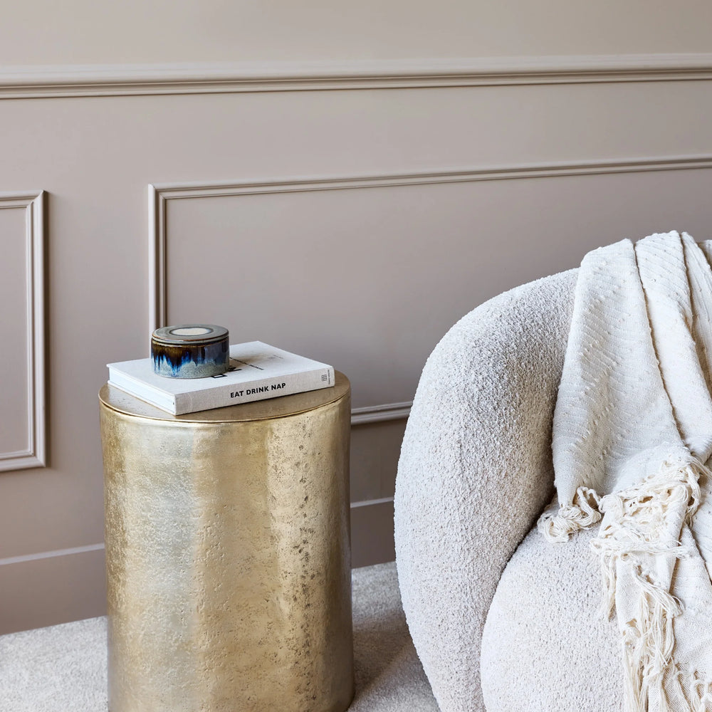 Gold metal side table beside a cozy cream armchair with a throw blanket.