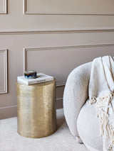 Gold metal side table beside a cozy cream armchair with a throw blanket.