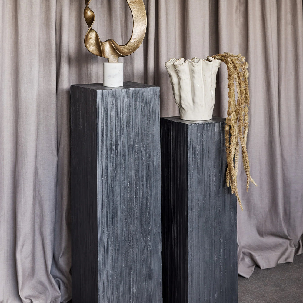 Dark wooden plinths with abstract sculpture and textured vase in front of curtains.
