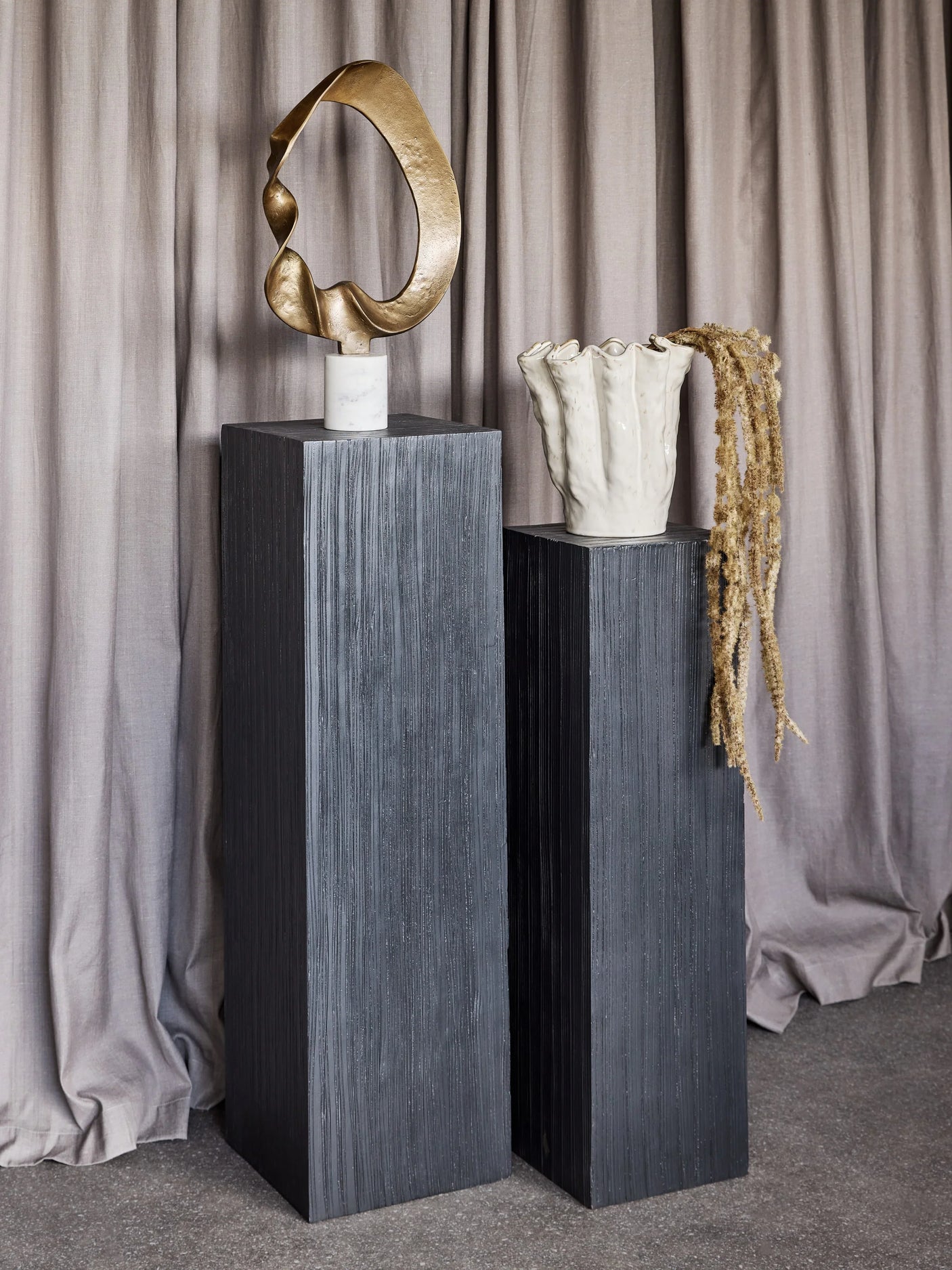 Dark wooden plinths with abstract sculpture and textured vase in front of curtains.