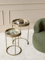 Golden side tables with jungle design, styled next to a green chair on beige carpet.