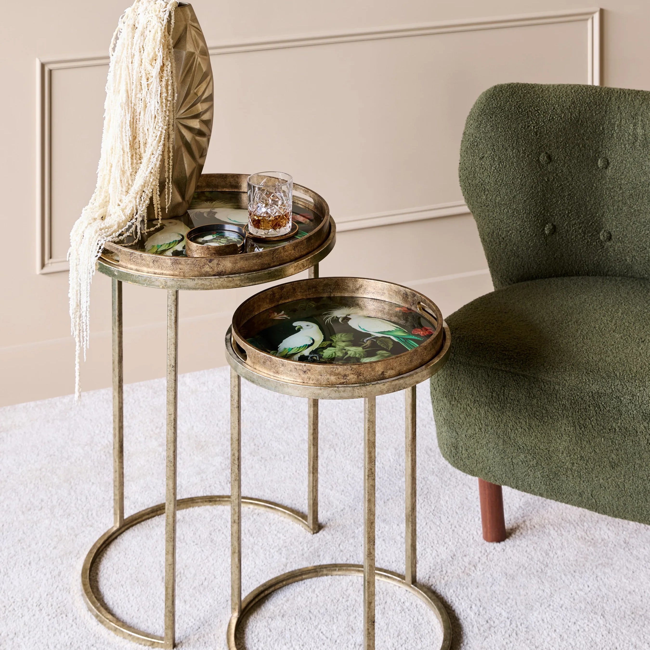 Gold side tables with tropical bird designs beside a green chair in a cozy room.