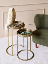 Gold side tables with tropical bird designs beside a green chair in a cozy room.