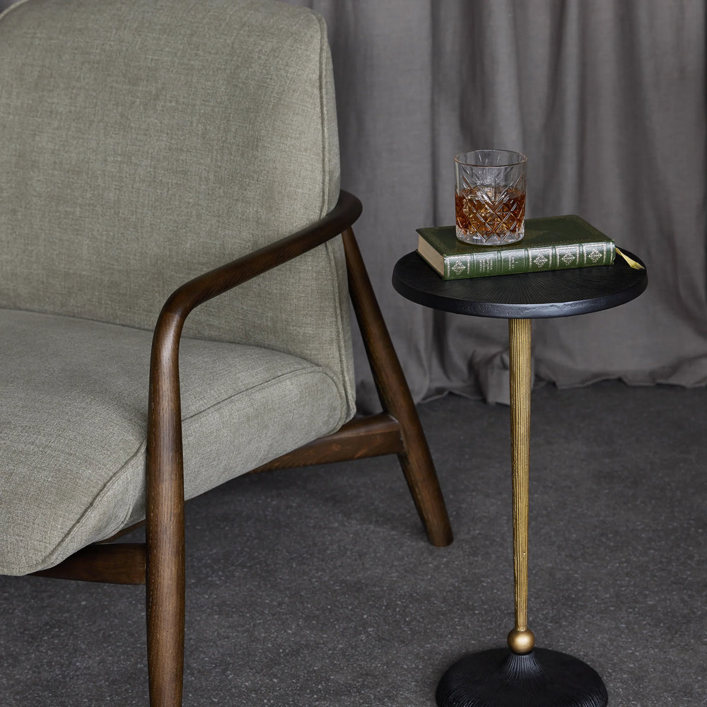 Black and gold round side table beside a grey armchair with a book and drink on top.