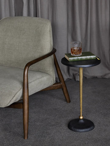 Black and gold round side table beside a grey armchair with a book and drink on top.