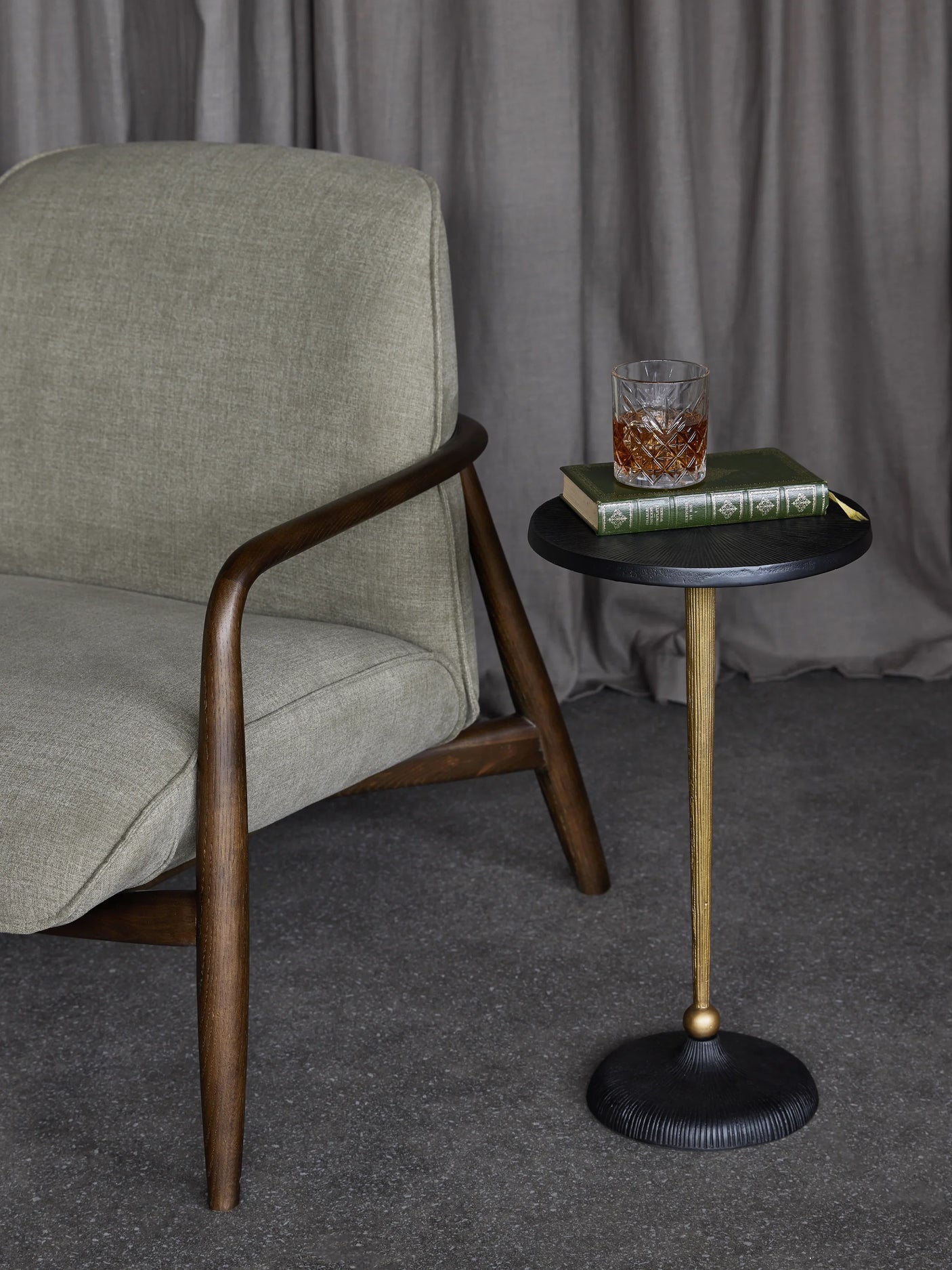 Black and gold round side table beside a grey armchair with a book and drink on top.