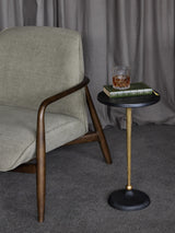 Black and gold round side table beside a grey armchair with a book and drink on top.